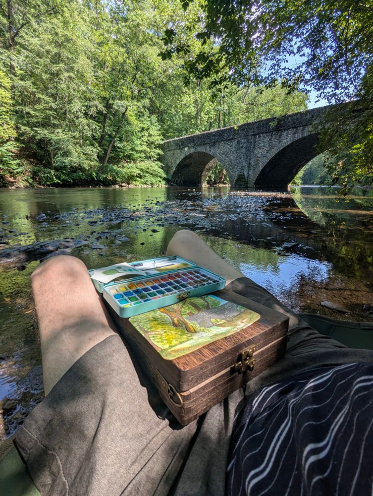 A photo of yours truly sitting by a riverbank near an old stone bridge, painting a scene.