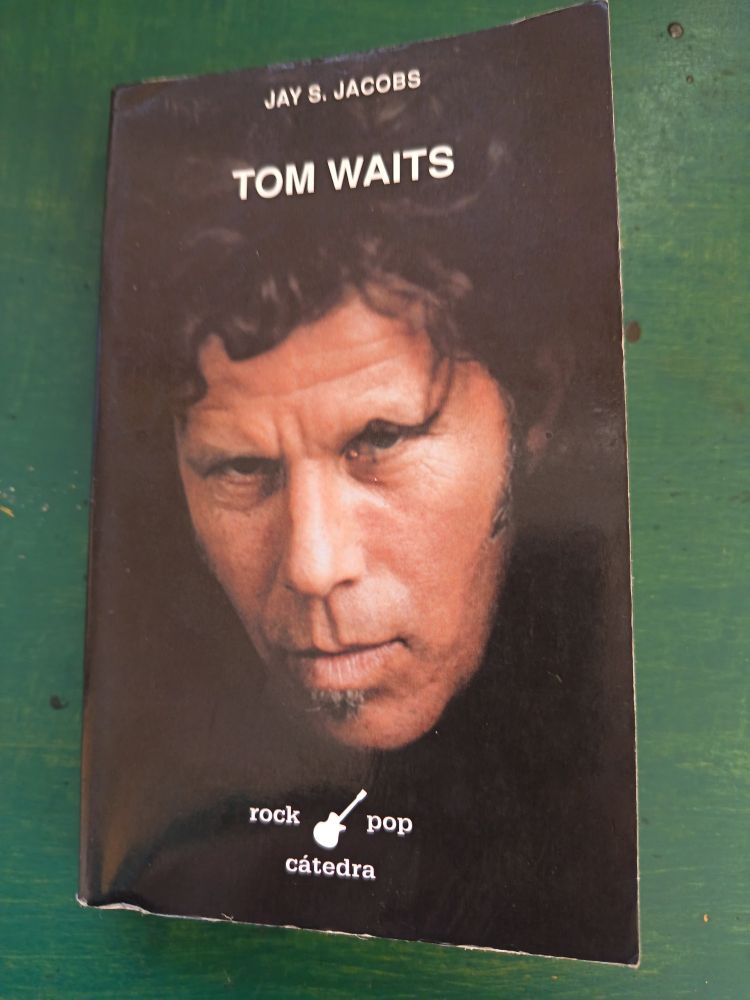 Picture of a book about Tom Waits which has a photograph of him against a black background on the cover. The book is on a green painted wooden table