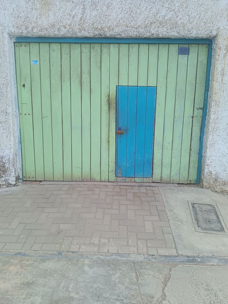 a turquoise door  inside pale green painted vertical wooden planks surround with turquoise border at top and right.. In the foreground is different textured and coloured brick and concrete ground with a utilities cover. Around the doorway is rough plastering painted white