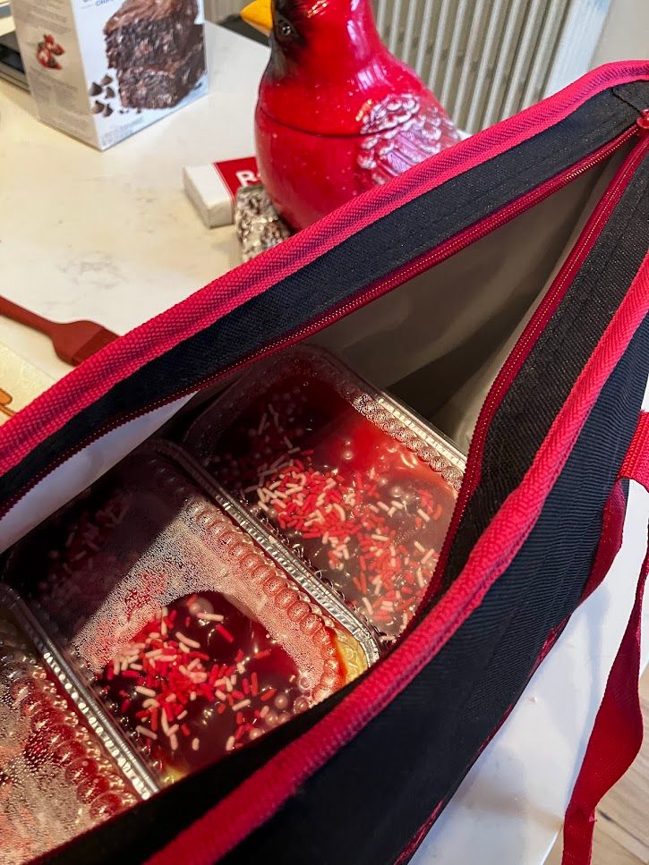 A photo of a kitchen counter with a red bag slightly unzipped. The camera is looking into the bag, and there appear to be a lot of cheesecakes hiding in it. There is a large red ceramic cardinal in the background, again, in profile to the left. There is also the edge of a brownie mix just peeking into the frame from the top left.