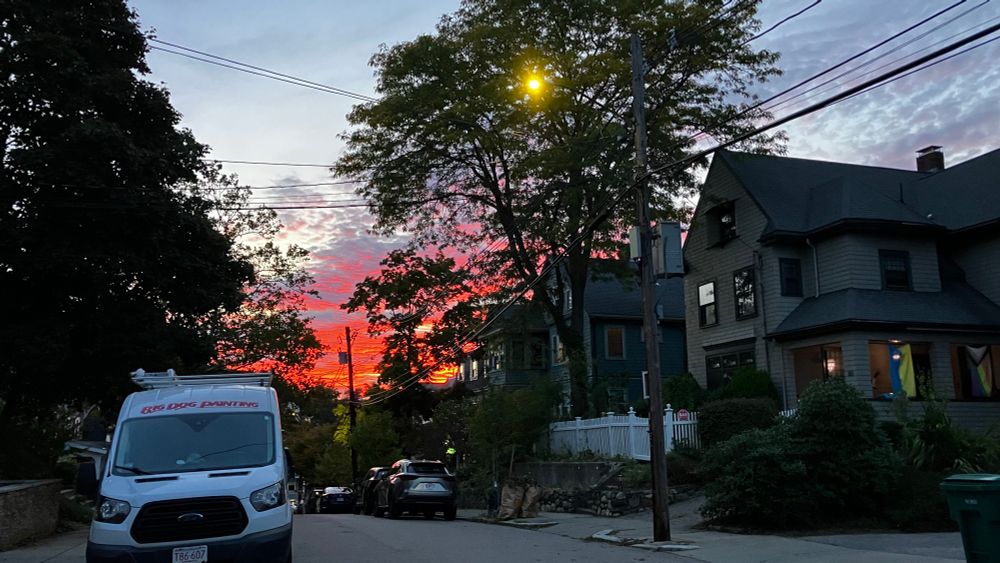 A dusk photo of a Boston neighborhood with a white work van on left of street, house on right, and a brilliant red and pink hued sunset in the sky behind a large hill 