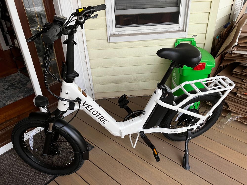 A photo of a porch with a small electric bike, colored white, with it giving a side profile to the camera. There is a window and a small green trash can behind the bike. A door in the upper left is ajar.
