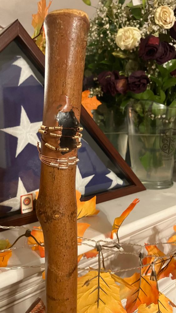 A close up digital photo of a wooden staff with a chunk of obsidian in it. There are copper wires wrapped around the stick and obsidian, and each copper wire has small coils of golden thread spaced out to either side of the obsidian. Below the obsidian wrap, another coil of copper but with silver coils is affixed tightly.
In the background is a showdown’s with an American flag