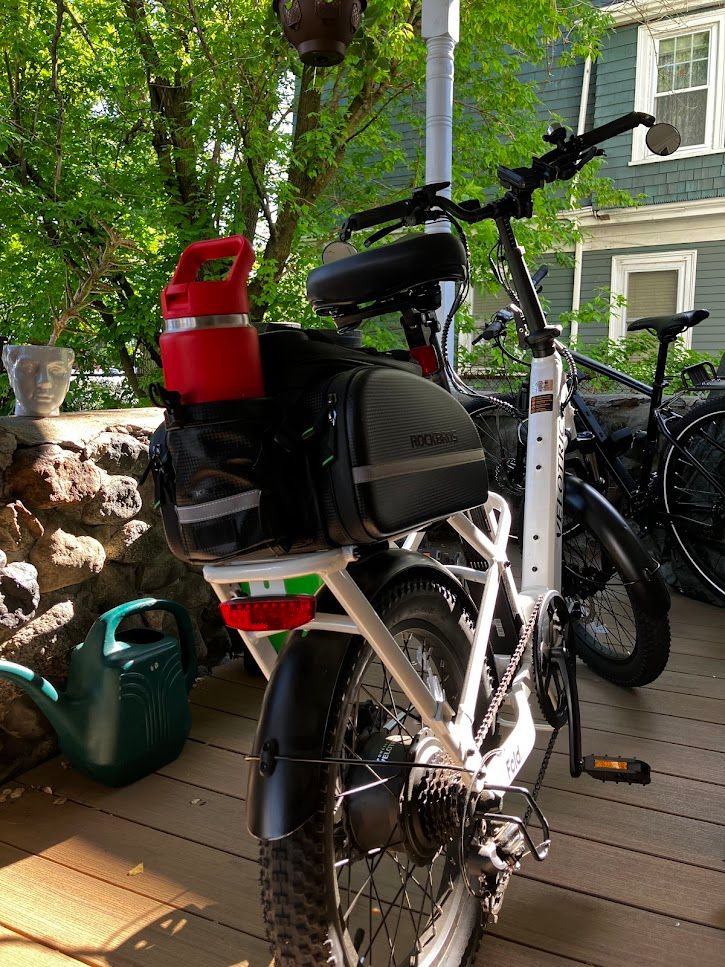 A rear view photo of a bike on a porch. It is white, and the view is from rear. There is a red water bottle ina black saddle bag. 