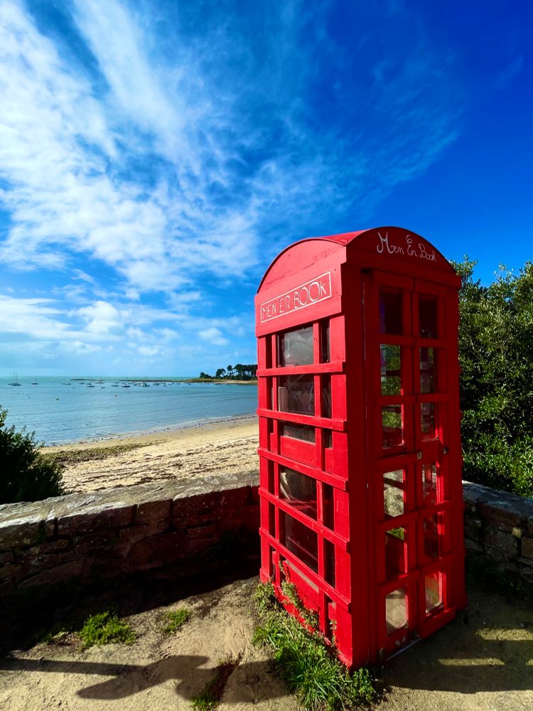 Une cabane à livre rouge dans une sorte de cabine téléphonique anglaise devant une plage de sable jaune et une mer bleu avec un ciel un peu nuageux 