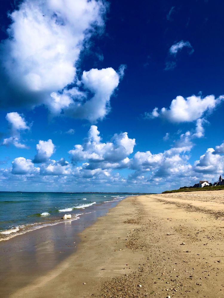 Plage de sable jaune avec mer à droite, qq maison a gauche ciel avec des gros nuages blancs epars