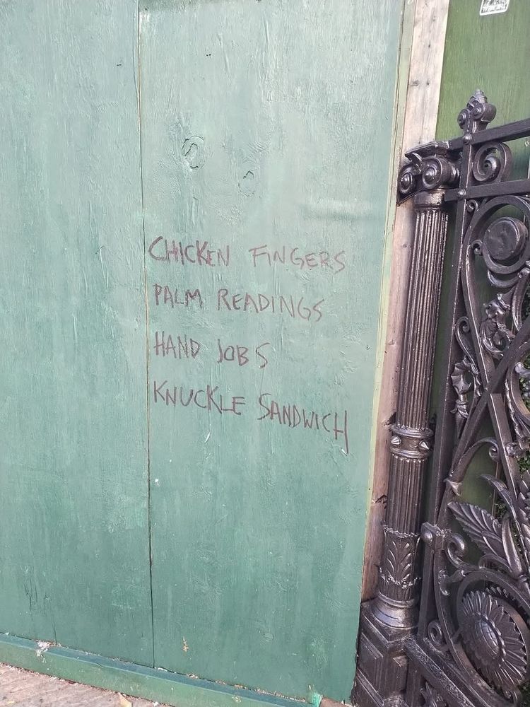 Green wall with written graffiti listing "chicken fingers, palm readings, hand jobs, knuckle sandwich" 