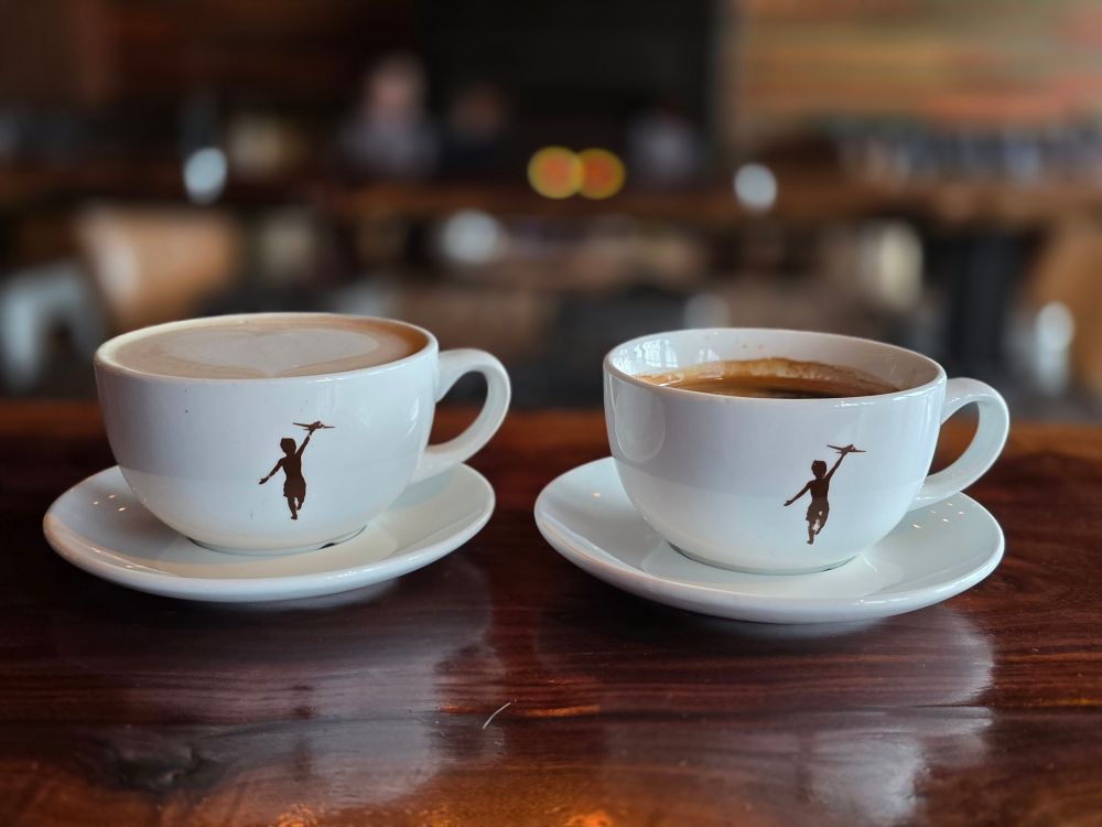 Two cups of coffee with a coffee shop sitting area in the background