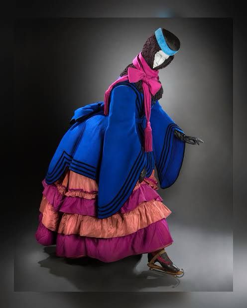 An action filled photograph of a mannequin posed in an1870s winter ice skating ensemble consisting of layered skirts, a blue wide sleeved mantle with a shawl and warm hat/scarf tied at the neck