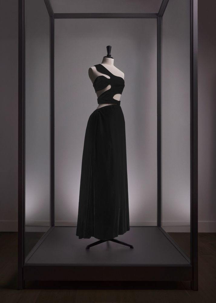 A black pleated evening dress with a variety of cut outs around the torso and a long draped and pleated skirt