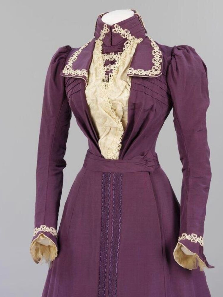 The top half of a purple ensemble showing a high collared bodice with long sleeves, trimmed with white lace and a white insert centre front, the top of the matching skirt just visible 