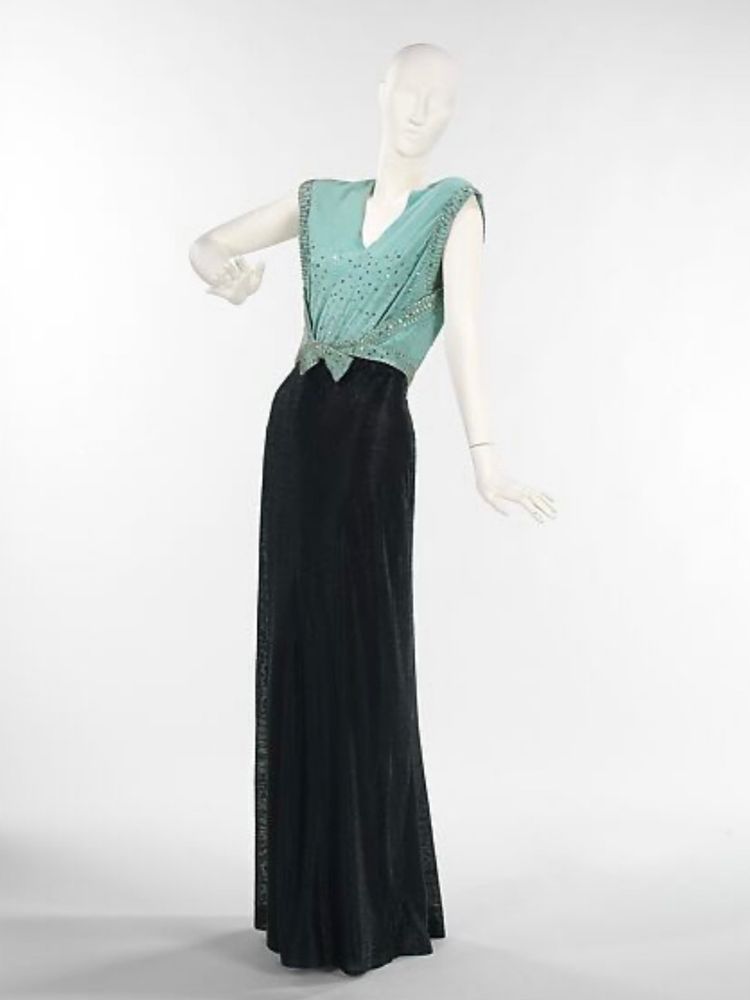 A mannequin wearing a long narrow sleeveless dress. It has a slim black skirt and a pale turquoise knotted bodice with a V neck. The bodice is sprinkled with rhinestones 