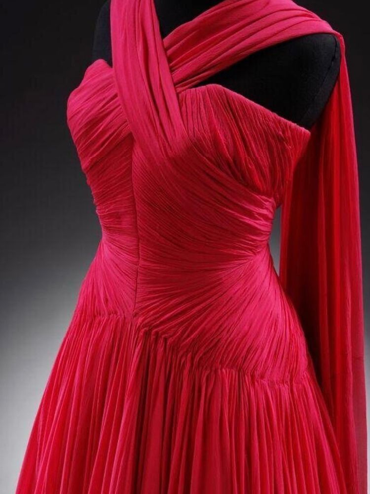 A photograph of the top half of a bright red sleeveless silk jersey evening dress with a complicated pleated design at the bodice fanning out into a softly draped skirt. It has a crossover integral scarf that falls centre back