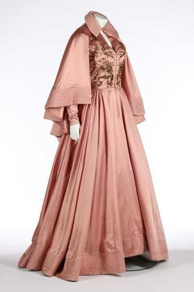 A three quarter view of a long pink silk satin dress with bands of stitching around the hem. The bodice is embellished with metallic embroidery. It has a matching shoulder cape