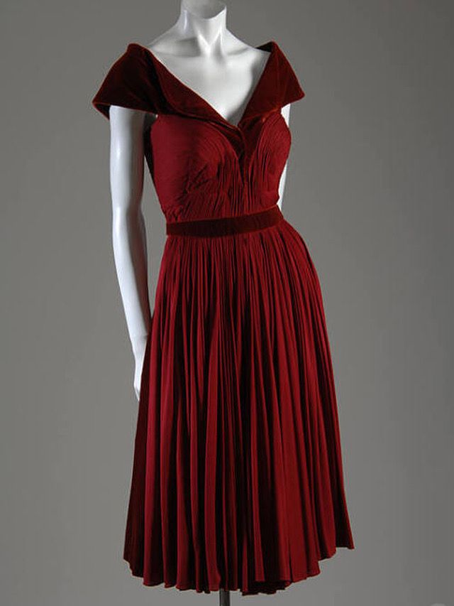 A sleeveless red pleated silk jersey cocktail dress. It has a red velvet collar that creates a V neck and sweeps over the shoulders with a matching red velvet belt