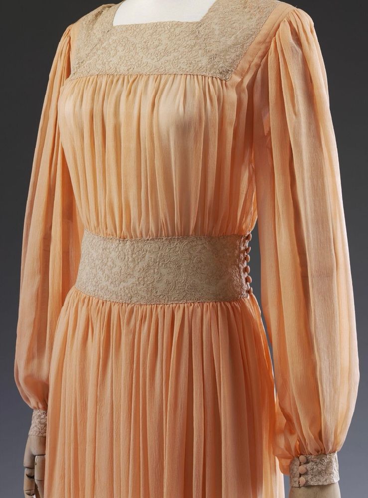 A photograph of the mid section of a 1940s peach silk tea gown showing the square neckline, long sleeves and lace inserts. The inserts decorate the neck, cuffs, waist and skirt