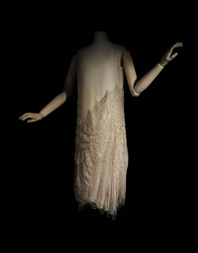 A mannequin posed before a shadowy background wearing a pale sleeveless dress, the bottom portion of the skirt decorated with a beaded cobweb design