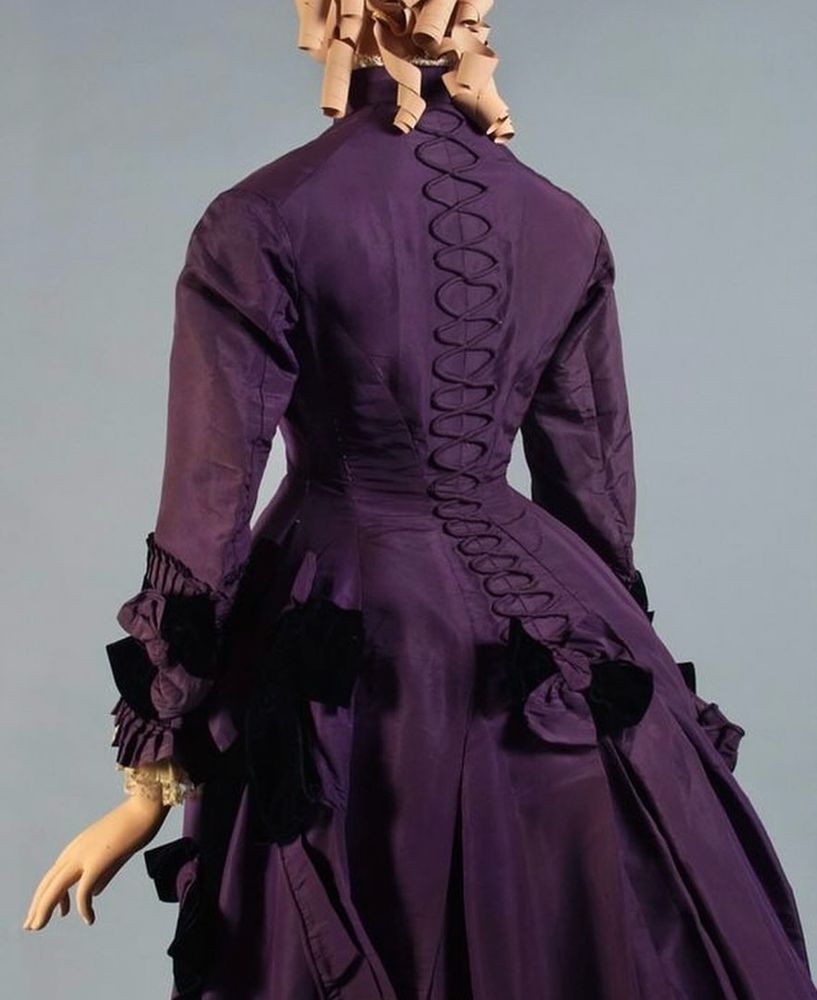Back view of a mannequin wearing a purple dress showing the top half of the bustle style gown. It has long sleeves with pleated cuffs, decorative lacing detail centre back and self coloured bows above the draped bustle skirt
