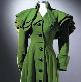 A long green wool coat with long sleeves and deep cuffs. It has a many tiered collar and large black buttons fastening centre front. Designed by Rene Hubert 