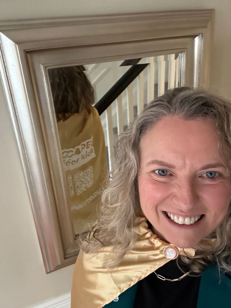 Woman with blond hair and black top wearing gold cape with “capes for kids” visible in mirror