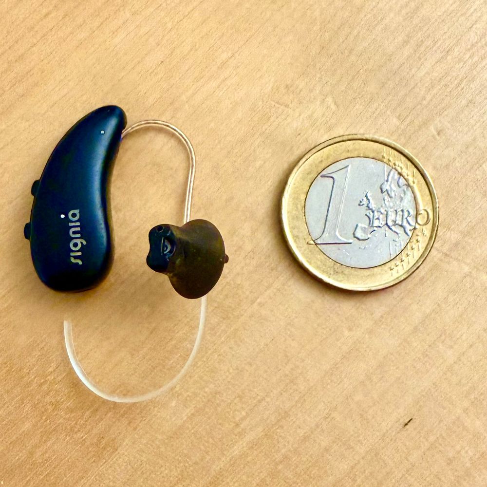 Ein schwarzes Hörgerät der Marke Signia liegt neben einem 1-Euro-Stück. Beide sind ähnlich gross mit Blick auf die Höhe.