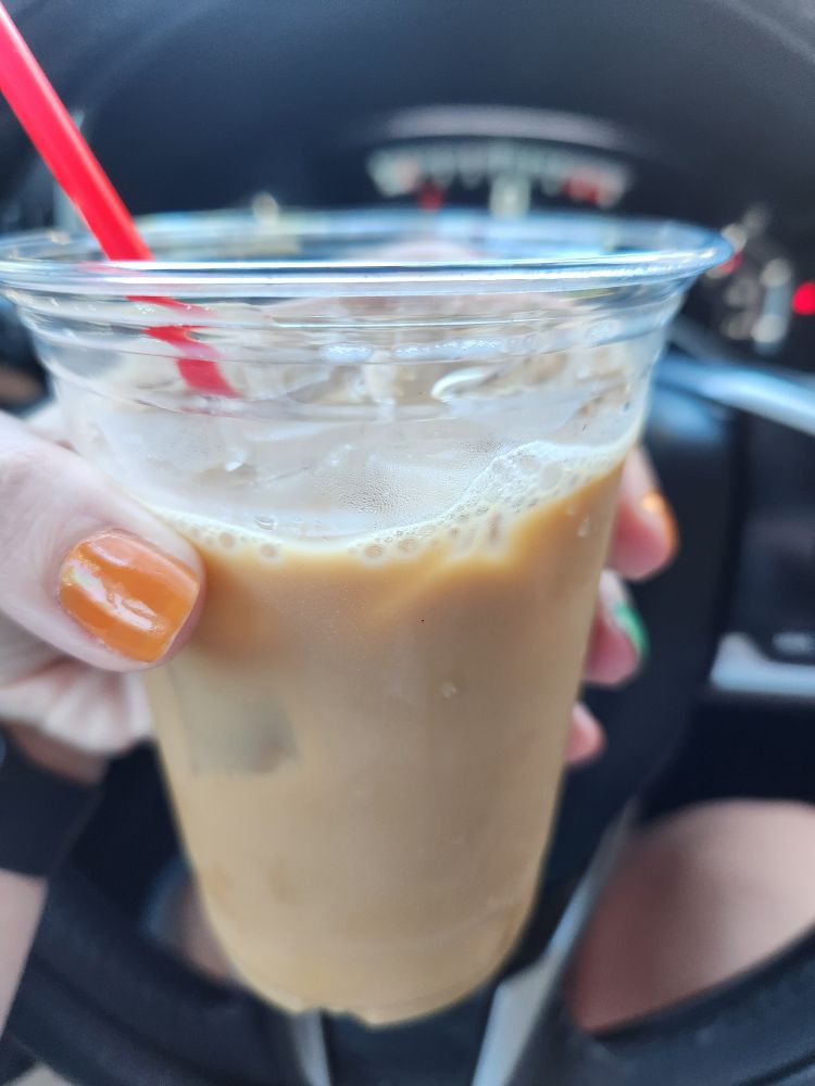 Woman's hand holding a 16oz cold brew drink with whole milk and caramel syrup.
Her nails are painted orange and green to match Taylor's Showgirl theme, thanks to her Swiftie kid.