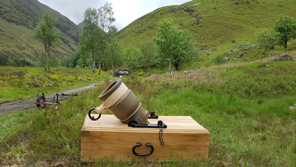 Replica of a bronze coehorn mortar on its wooden base sitting in mountain pass of Glenshiel