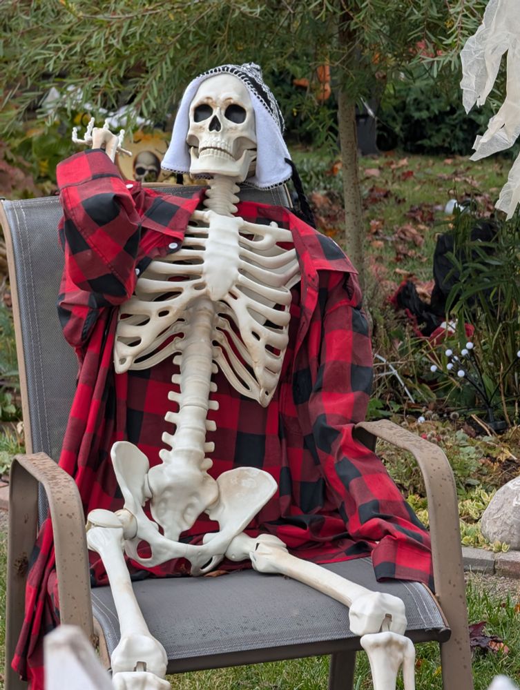 A neighborhood skeleton dressed in red flannel and a touque as we call them in Canada sitting around his campfire.
