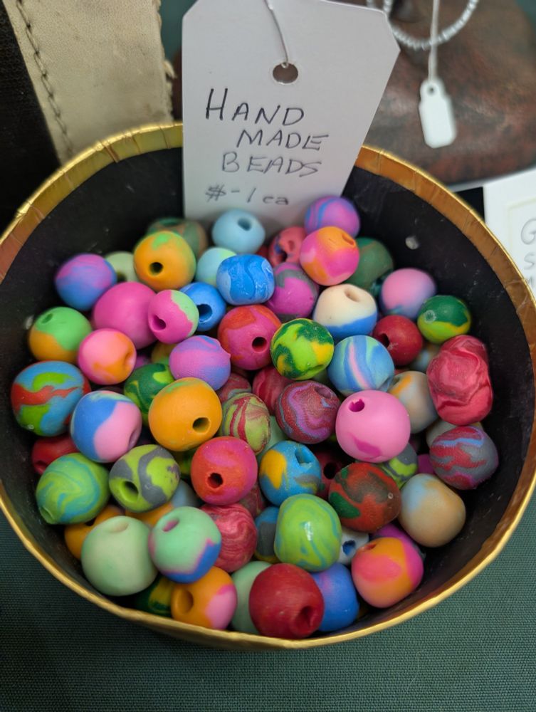 A gold circular container holding multiple round rainbow colored polymer beads. A tag sign sticking out of the container reads Handmade beads $1 each. 