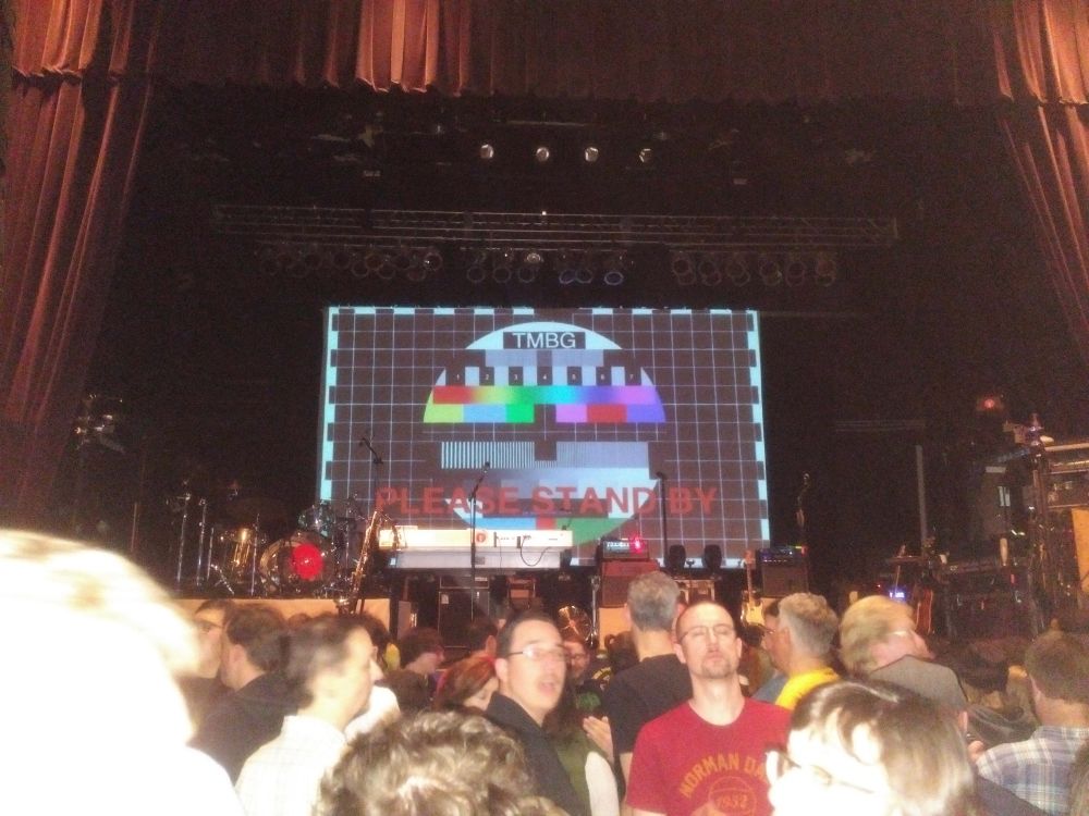 A small concert auditorium. A crowd of people in the foreground facing in various directions. On the stage stand a drum kit and some microphone stands and amplifiers.
The background of the stage is dominated by a projector screen with the word "TMBG" at the top and the phrase "please stand by" at the bottom. The middle of the screen shows a circle containing rows of variously spaced blocks of color, black, gray, and rainbow gradients in the style of a television test pattern.
The house lights are still on, the concert has not yet started. 