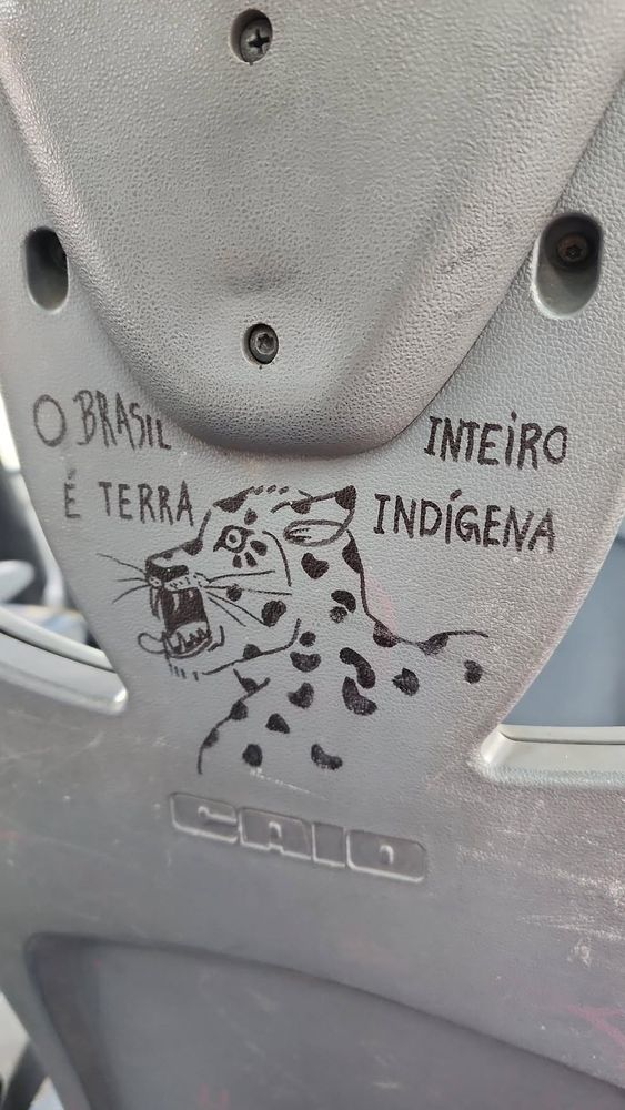 parte de trás do assento de um ônibus com escritas em caneta preta "O Brasil inteiro é terra indígena" e o desenho de uma onça com a boca aberta, dentes e língua a mostra