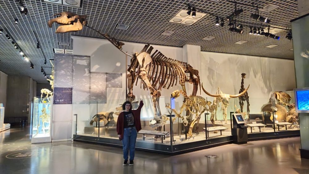 Eu (mulher de 30 anos, cabelos encaracolados, usando calça jeans, blusa preta e um cardigan vinho) na frente de réplicas de ossos de dinossauros, um deles medindo mais de 3 metros de altura