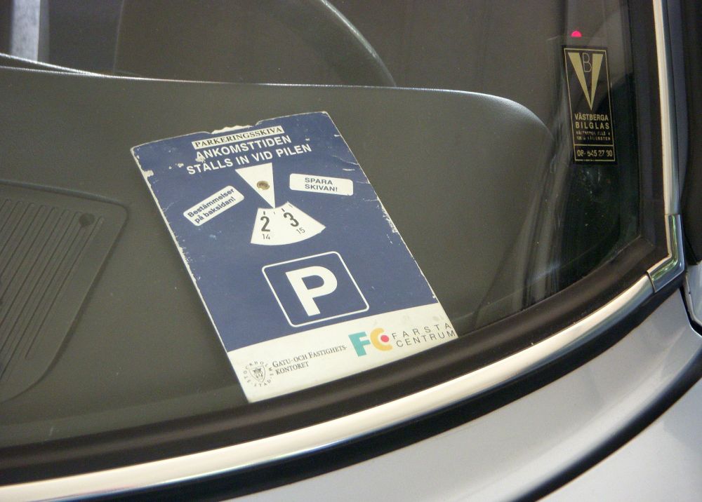 Photograph of a European 'parking disc' on the dashboard of a car. The parking disc is a blue rectangle, with a white "P" parking symbol printed on it and a white arrow, and a white rotating disk that has times printed on it. 