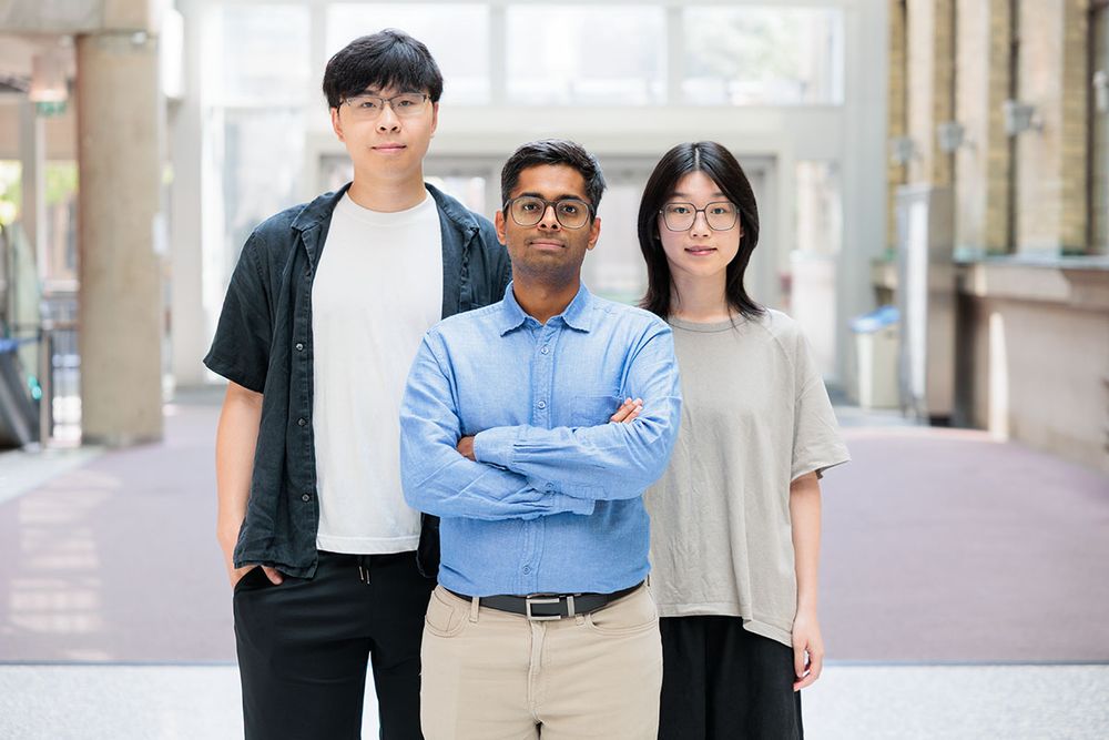 Three people stand facing the camera with neutral facial expressions in a light-filled atrium. The man in the middle has his arms crossed.