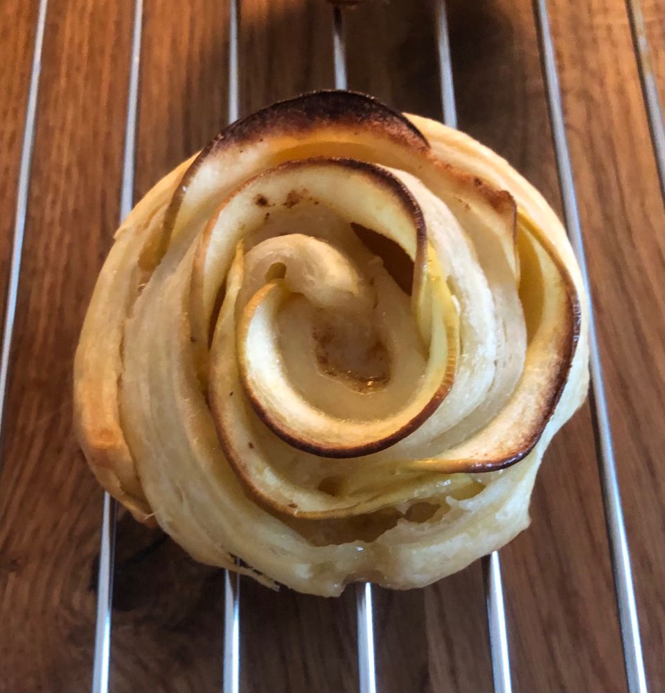 Das Bild zeigt einen goldbraun gebackenen Apfelstrudel, der wie eine Rose geformt ist und auf einem Gitterrost liegt.


Das Bild zeigt einen einzelnen, gebackenen Apfelstrudel in der Form einer Rose. Er liegt auf einem metallenen Abkühlgitter, das auf einer hölzernen Unterlage ruht. Der Strudel ist goldbraun und leicht karamellisiert an den Spitzen der Apfelscheiben. Die Apfelscheiben sind in konzentrischen Kreisen um einen Kern aus Apfelmus oder ähnlichem angeordnet, wodurch die Rosenform entsteht.  Der Teig ist knusprig und leicht gebräunt. Das Holz der Unterlage ist warm und hellbraun. Die Atmosphäre ist warm und gemütlich, die Stimmung ist appetitanregend und beruhigend. Das Foto ist von oben aufgenommen, wobei der Strudel die volle Aufmerksamkeit auf sich zieht. Die Beleuchtung ist gleichmäßig und lässt die Details des Gebäcks gut erkennen.
