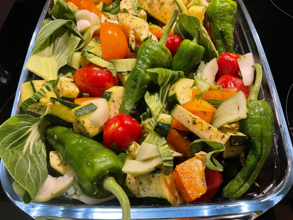Das Bild zeigt eine Auflaufform aus Glas, gefüllt mit verschiedenen, gewürzten und in mundgerechte Stücke geschnittenen Gemüsesorten.  Man erkennt unter anderem Zucchini, Tomaten (rot und gelb), Paprika (grün), Zwiebeln und Pak Choi. Das Gemüse scheint mit Kräutern und Gewürzen gewürzt zu sein und bereit zum Backen oder Braten zu sein.
