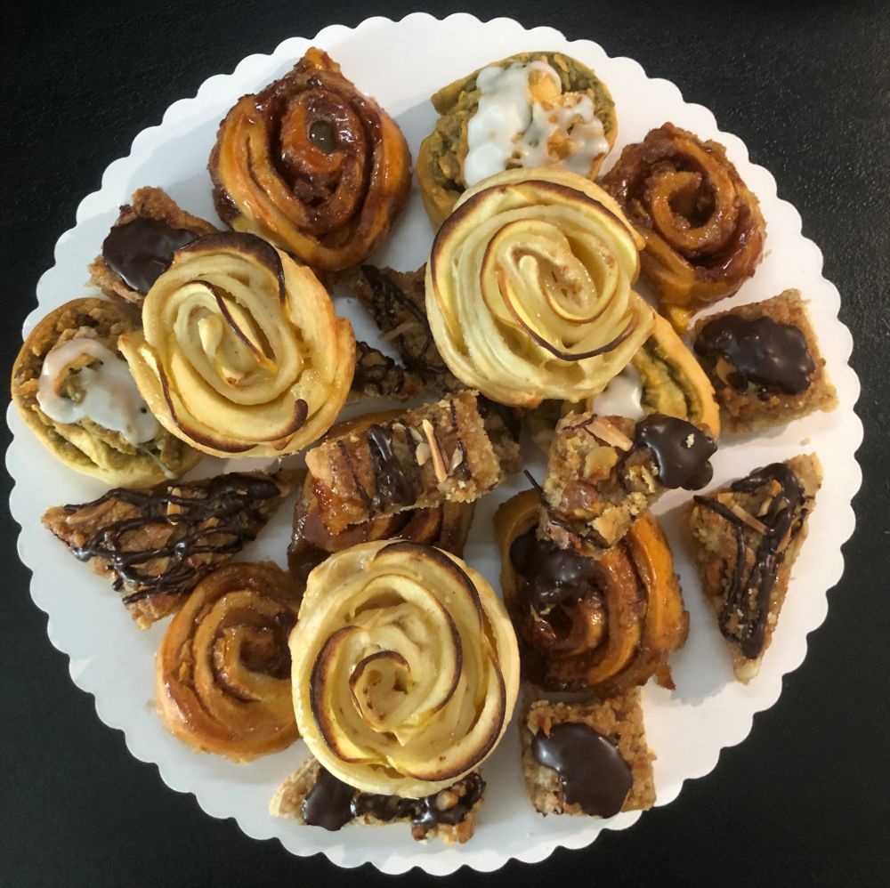 Das Bild zeigt eine Auswahl verschiedener süßer Backwaren auf einem weißen Teller. Es sind Zimtschnecken, Apfelrosen und Gebäckstücke mit Schokoladenüberzug zu sehen. Die Backwaren sehen frisch gebacken und appetitlich aus.  Sie scheinen aus Blätterteig oder ähnlichem Gebäck hergestellt zu sein und sind mit verschiedenen Toppings wie Zimt, Zuckerguss und Schokolade verziert.
