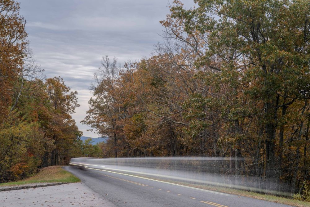The Blue Ridge Parkway near Roanoke, VA. The trees are showing their fall colors of red, orange, yellow, and green as a car rushes past the display. The photo was taken with a long exposure, elongating the car into a white and red blur on the Parkway.