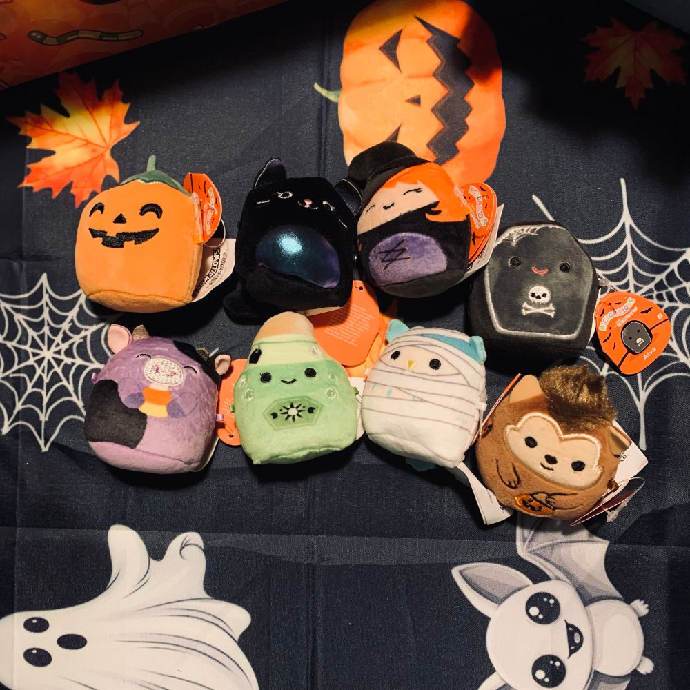 Eight Halloween Micromallows for the squishmallow Halloween Advent calendar where the one on the left is a 2 inch pumpkin and in the middle there is a 2 inch black cat, a 2inch witch, a 2inch coffin, a 2inch purple cow eating a candy corn, a 2inch green potion bottle, a blue owl dressed as a mummy and the one last one is a 2inch werewolf holding a pumpkin trick a treat bag.