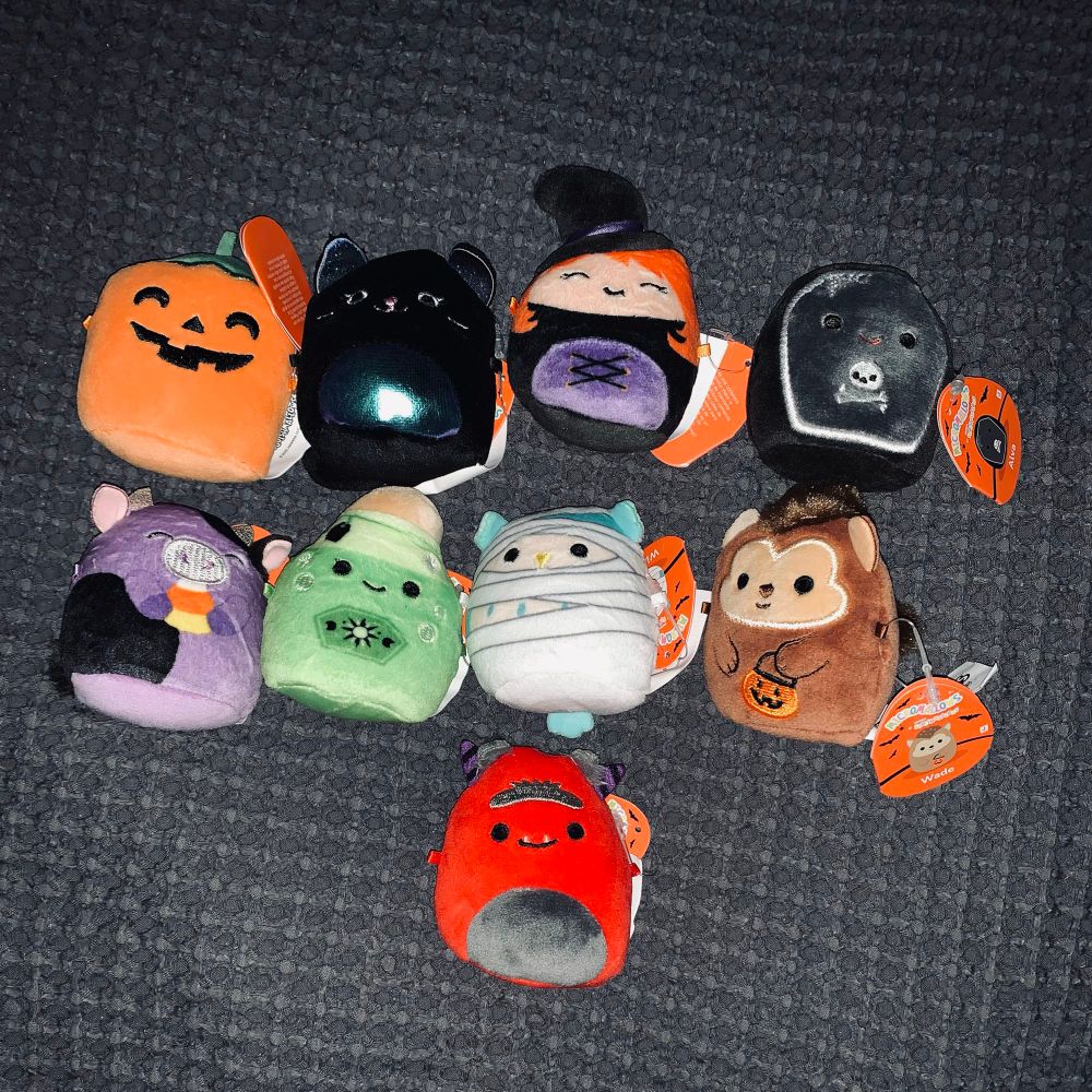 Nine Halloween Micromallows for the squishmallow Halloween Advent calendar where the one on the left is a 2 inch pumpkin and in the middle there is a 2 inch black cat, a 2inch witch, a 2inch coffin, a 2inch purple cow eating a candy corn, a 2inch green potion bottle, a blue owl dressed as a mummy, a 2inch werewolf holding a pumpkin trick a treat bag and the one last one is a 2inch red devil with a monobrow.