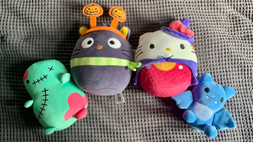 Four plushies; from left to right there is a chubby zombie, Chococat squishmallow wearing a pumpkin antenna headband, hello kitty squishmallow dressed as a witch, an a little blue bat.