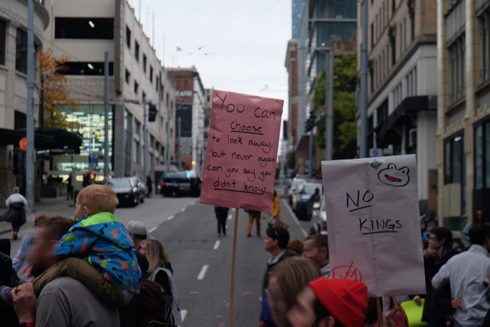 As the protest crosses the road, a sign reads "You can *choose* to look away, but never again can you say you *don't know.*"