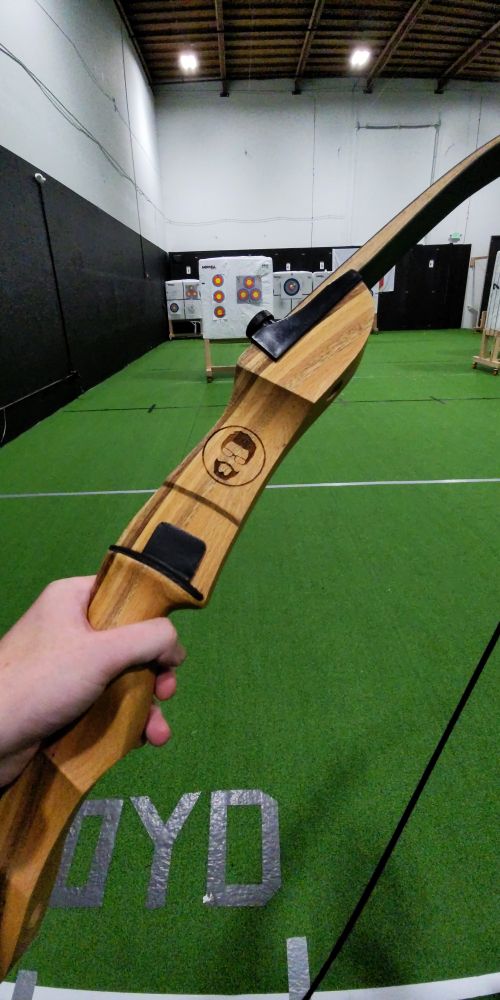 deusjz holds a recurve bow in his left hand in front of an indoor range. the riser is etched with his logo in the wood above the shelf.