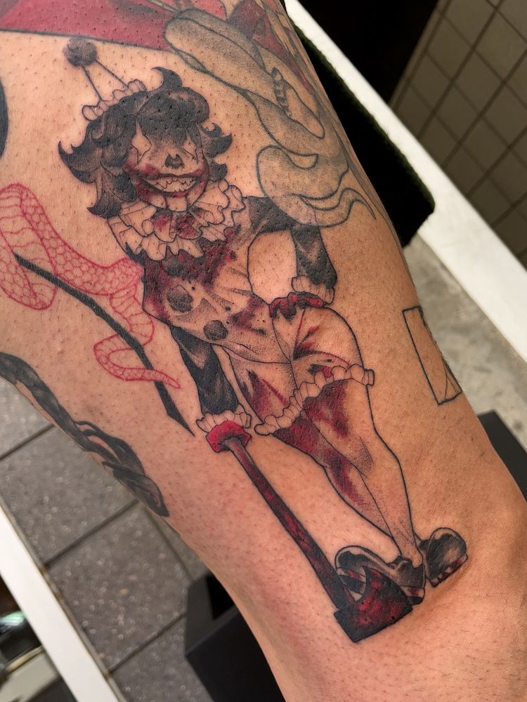 A tattoo depicting a femme clown character with a large, toothy grin. She’s leaning on a bloody axe, and her outfit is covered in blood.