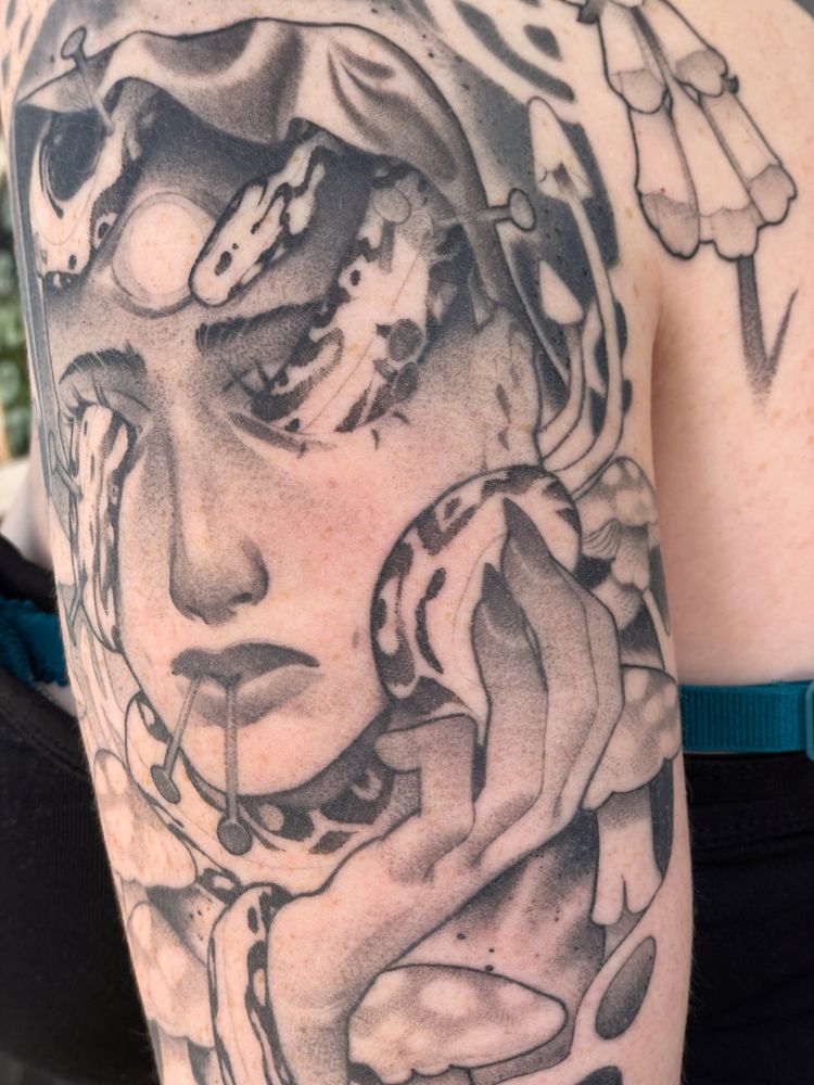 A tattoo depicting the face of an androgynous character with a snake moving through their eye sockets. Throughout the tattoo are mushrooms, plants, and other fantastic and floral imagery.