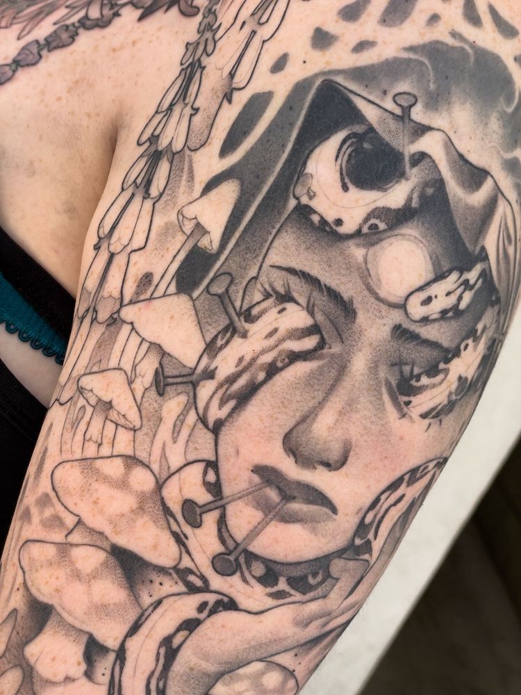 A tattoo depicting the face of an androgynous character with a snake moving through their eye sockets. Throughout the tattoo are mushrooms, plants, and other fantastic and floral imagery.