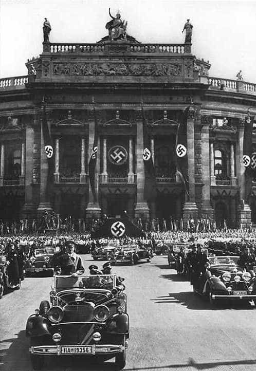 A photograph of Adolf Hitler in a motorcade with swastika flags and crowds