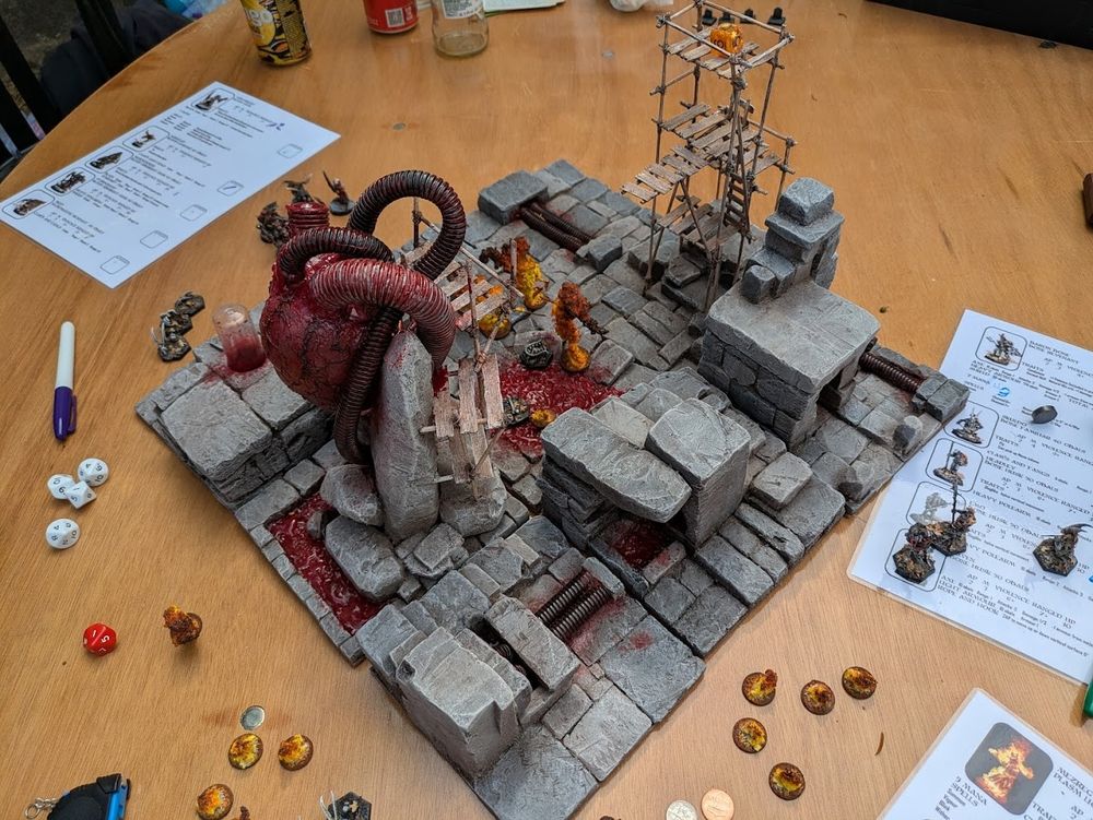 A game of the skirmish game Necropolis in progress on a small board that features a terrain piece in the shape of an enormous heart pumbing blood through pipes that run across the board. 