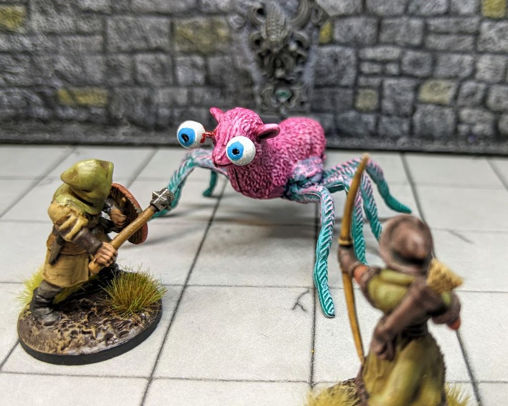 A scene with miniature figures: two fantasy adventurers confront a hideous creature, part lamb, part spider. 