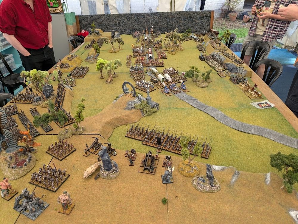 A massive game of Warhammer Fantasy Battle about to start. Orcs and undead prepare to ambush a caravan of wagons and troops. There are hundreds if not thousands of figures on the table. 